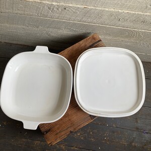 May include: White ceramic baking dish with a matching lid. The dish has a square shape with rounded corners and two handles on the sides.