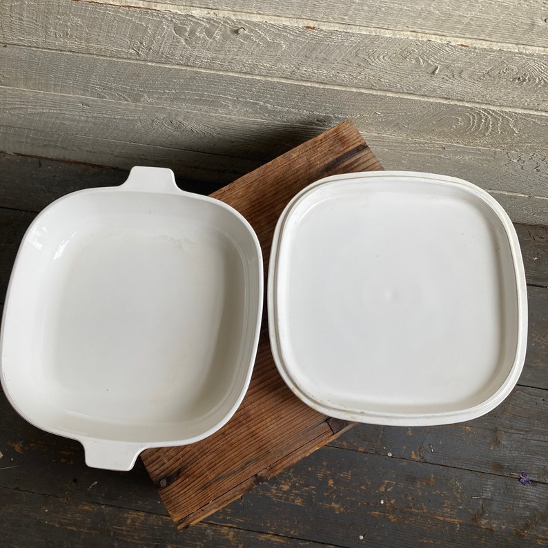 May include: White ceramic baking dish with a matching lid. The dish has a square shape with rounded corners and two handles on the sides.