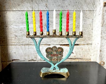 May include: A brass menorah with nine colourful candles. The candles are green, yellow, red, blue, white, and yellow. The menorah has a decorative emblem in the centre and sits on a black surface.