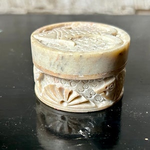 May include: A small, round, stone trinket box with a carved design of fish and a fan. The box is made of light grey stone and has a lid.