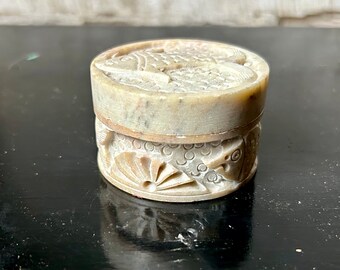 May include: A small, round, stone trinket box with a carved design of fish and a fan. The box is made of light gray stone and has a lid.