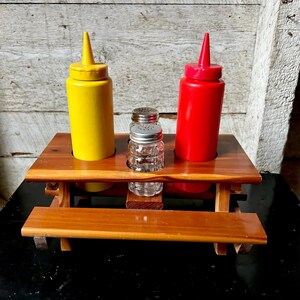 Pode incluir: Um suporte de condimentos em forma de mesa de piquenique de madeira com uma garrafa de mostarda amarela, uma garrafa de ketchup vermelha e um saleiro de vidro transparente. As garrafas de mostarda e ketchup têm pontas.