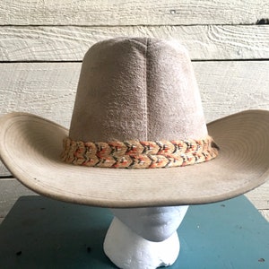 May include: A light brown cowboy hat with a woven band featuring orange, black, and white accents.