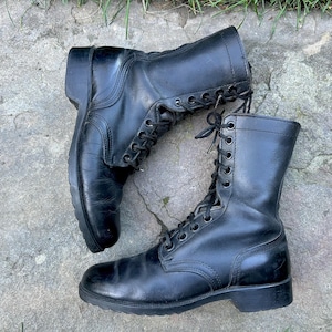Puede incluir: Un par de botas de combate de cuero negro con diseño de caña alta, cordones frontales, ojales metálicos, cordones negros, punta redondeada y tacón bajo. Las botas tienen un acabado liso y una caña estructurada que llega sobre el tobillo.