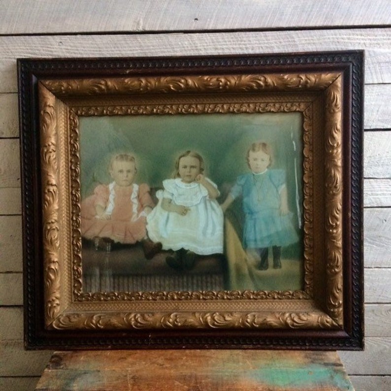 May include: An antique picture frame with a gold-colored ornate border and a dark brown wood exterior. The frame contains a faded photograph of three young girls in vintage clothing.