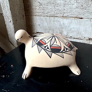 May include: A ceramic turtle figurine with a cream-colored body and black, red, and white geometric designs on its shell. The turtle has four stubby legs and a small head. The figurine is displayed on a black surface.