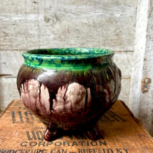 May include: A small, round ceramic planter with a dark brown base and a green rim. The planter has three small feet and a dripped glaze effect in shades of brown and cream. The planter is sitting on a wooden crate.