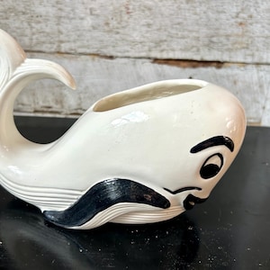 May include: A white ceramic whale-shaped planter with a black mustache and eye details. The whale has a curved tail and a wide opening for plants. The planter is on a black surface.