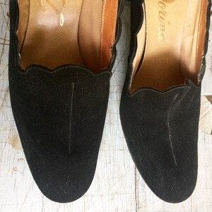 May include: A pair of black suede heeled shoes with scalloped edges and the word "Divina" written on the inside of each shoe.