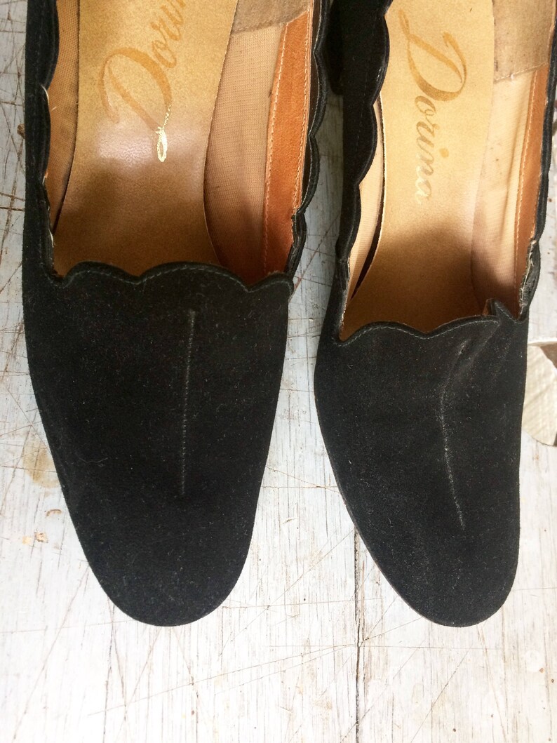 May include: A pair of black suede heeled shoes with scalloped edges and the word "Divina" written on the inside of each shoe.