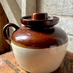 May include: A two-tone stoneware jug with a lid. The upper portion is a rich brown, transitioning to a cream-colored base. It features a handle and a matching brown lid with a central knob. The jug is likely vintage or antique.