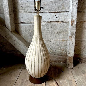 May include: A vintage table lamp with a cream-colored, ribbed ceramic body and a dark brown base. The lamp has a classic teardrop shape and a brass neck, topped with a light bulb. The lamp's cord is visible.
