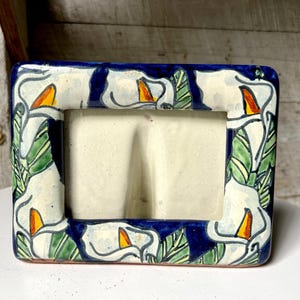 Könnte beinhalten: Rechteckiger Keramik-Bilderrahmen mit weißem Innenraum. Der Rahmen ist mit weißen Calla-Lilien mit gelben Zentren und grünen Blättern vor einem dunkelblauen Hintergrund verziert. Der Rahmen steht auf einer weißen Oberfläche.