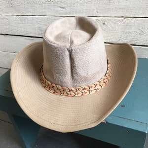 May include: A light brown suede cowboy hat with a braided band featuring orange and black accents. The hat is sitting on a teal blue wooden stool.