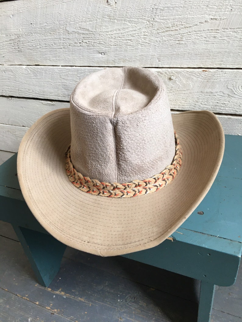 May include: A light brown suede cowboy hat with a braided band featuring orange and black accents. The hat is sitting on a teal blue wooden stool.