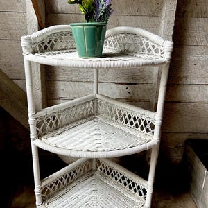 Op de afbeelding: Witte hoekplank van riet met drie driehoekige planken. Een groene keramische pot met roze en paarse bloemen staat op de bovenste plank. De plank is gemaakt van geweven riet en heeft een vintage uitstraling.