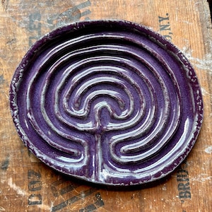 May include: A round, deep purple ceramic dish featuring a raised, swirling labyrinth pattern in a lighter, metallic-toned glaze. The surface has a glossy finish with visible texture along the edges and a continuous, winding path design.