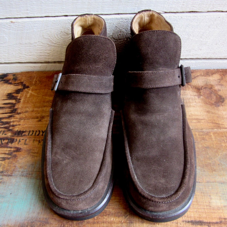 May include: A pair of brown suede ankle boots with a buckle strap. The boots have a rounded toe and a low heel.