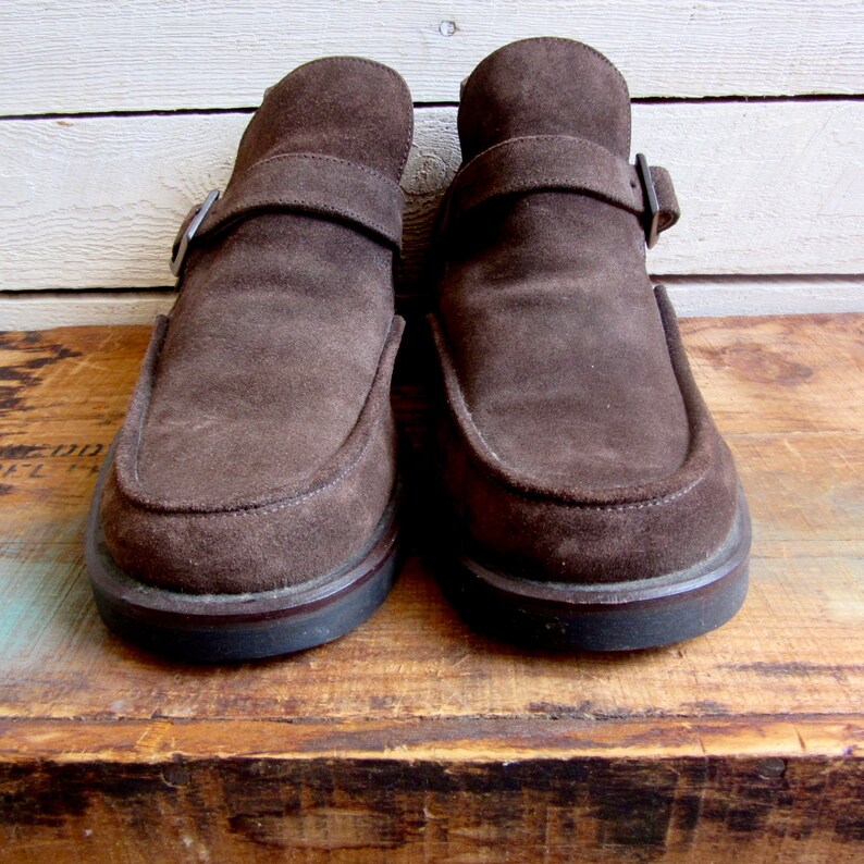 May include: A pair of brown suede loafers with a buckle strap. The loafers have a dark brown sole and are sitting on a wooden surface.