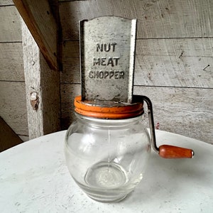 Op de afbeelding: Vintage glazen pot met een oranje deksel en een metalen noten- en vleeshakker eraan bevestigd. De hakmachine heeft een houten handvat en een metalen plaat met de tekst "Nut Meat Chopper".