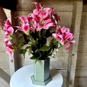Könnte beinhalten: Ein Strauß rosa Lilien in einer grünen sechseckigen Vase. Die Vase steht auf einem weißen Tisch. Die Lilien sind in voller Blüte und haben ein zartes, weibliches Aussehen.