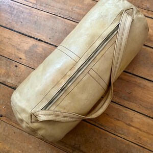 May include: A cylindrical, light tan leather bag with a zipper closure and a handle. The bag has visible stitching and a worn appearance, suggesting it is vintage or has been well-used. The bag is lying on a wooden surface.