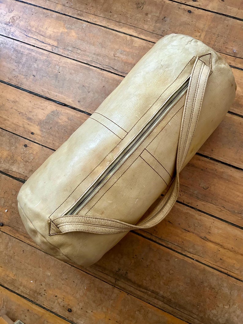 May include: A cylindrical, light tan leather bag with a zipper closure and a handle. The bag has visible stitching and a worn appearance, suggesting it is vintage or has been well-used. The bag is lying on a wooden surface.
