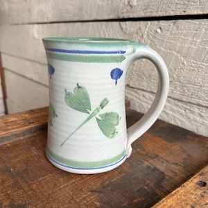 May include: White ceramic mug with a dragonfly design. The mug has a green and blue colour scheme, with a dragonfly illustration on the side. The mug has a handle and is suitable for hot or cold drinks.