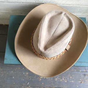 May include: A tan suede cowboy hat with a braided leather band. The hat is sitting on a blue surface.
