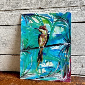 May include: An acrylic painting of a bird perched on a branch, set against a backdrop of turquoise and blue foliage. The bird has a red head, yellow and brown body, and a long, dark tail. The artwork is on a rectangular canvas.