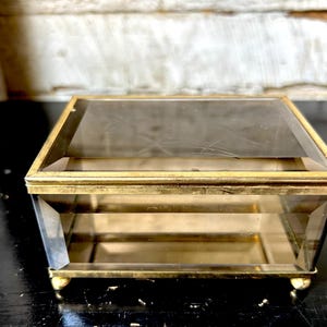 May include: A rectangular glass jewelry box with a brass metal frame. The box features beveled edges, a hinged lid with a faint etched star pattern on the top surface, and four small rounded brass feet at the base of the container.