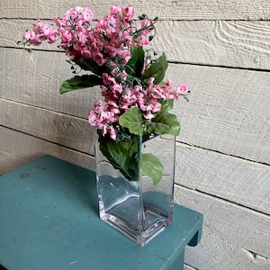 Può includere: Un vaso di vetro trasparente con base quadrata contiene un mazzo di fiori rosa con foglie verdi.