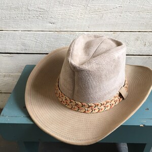 May include: A light brown suede cowboy hat with a braided band featuring orange and brown accents. The hat is sitting on a blue surface.