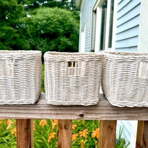 May include: Three white wicker baskets with a woven design. The baskets are sitting on a wooden railing.