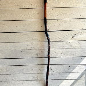 Puede incluir: Un bastón de madera con una mancha marrón oscura y un diseño en espiral negro. El bastón tiene una parte superior redondeada y una punta de goma negra.