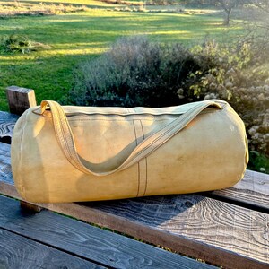 May include: A cylindrical, tan-colored duffel bag with a shoulder strap rests on a wooden bench. The bag has a zipper closure and vertical black stitching details. The background features a green field and sunlight.