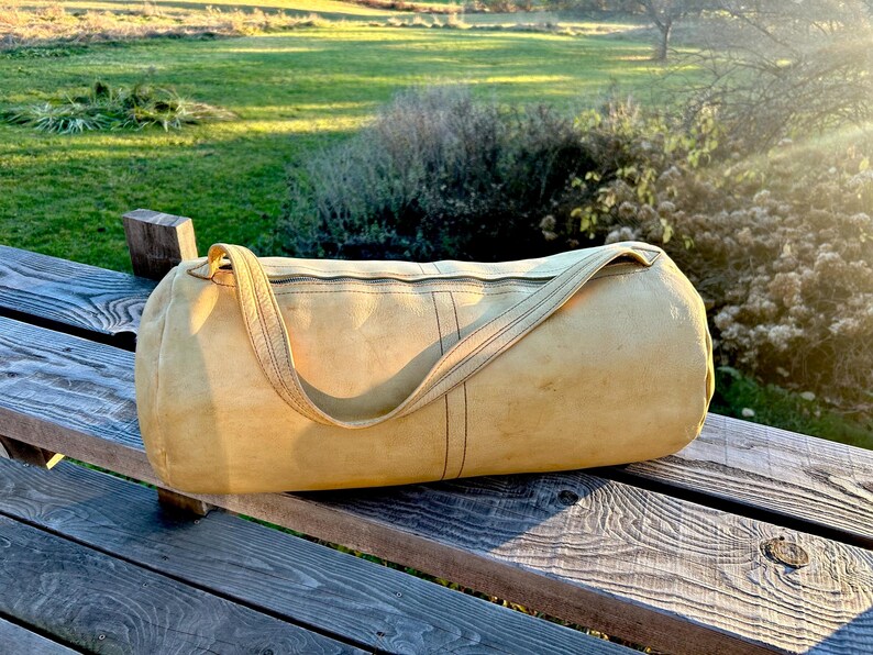 May include: A cylindrical, tan-colored duffel bag with a shoulder strap rests on a wooden bench. The bag has a zipper closure and vertical black stitching details. The background features a green field and sunlight.