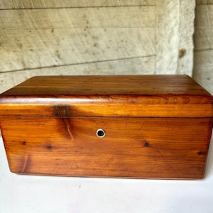 May include: A rectangular wooden box with a warm brown finish and visible wood grain patterns. The box features a smooth, flat lid and a small, circular metal keyhole centered on the front panel, showcasing a simple and functional design.