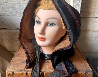Puede incluir: Una capucha negra vintage con ribete burdeos y un cuello a juego, exhibida en una cabeza de maniquí. La capucha está hecha de una tela brillante y tiene un forro marrón. La cabeza del maniquí tiene ojos azules y labios rojos. La capucha descansa sobre una caja de madera.