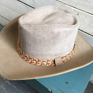 May include: A light brown suede cowboy hat with a braided band featuring orange and brown accents.