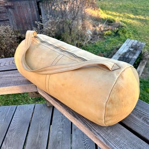 May include: A cylindrical, tan leather duffel bag with a top zipper closure and a single handle. The bag is made of a soft, worn leather and sits on a wooden surface. The bag is designed for travel or carrying sports equipment.