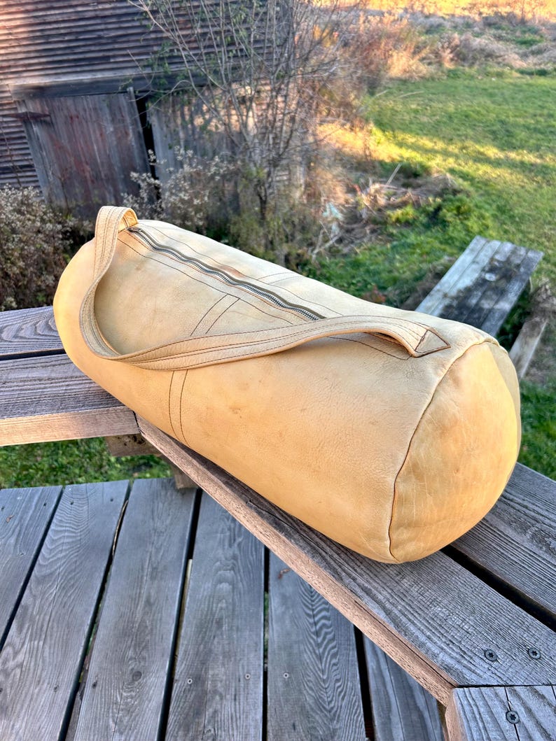 May include: A cylindrical, tan leather duffel bag with a top zipper closure and a single handle. The bag is made of a soft, worn leather and sits on a wooden surface. The bag is designed for travel or carrying sports equipment.
