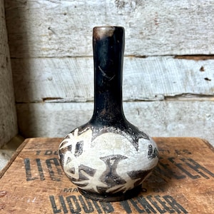 May include: A small ceramic vase with a rounded base and a long neck. The vase has a black top and a white body with a brown floral pattern. It rests on a wooden crate with the text "LIQUID VENEER CORPORATION". The vase is approximately 15 cm tall.