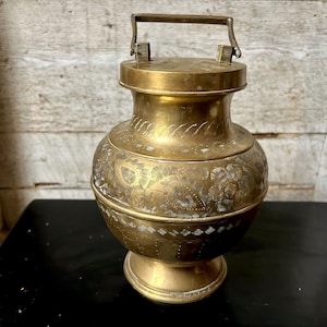 Puede incluir: Contenedor de latón antiguo con asa y tapa. El recipiente presenta patrones florales y geométricos grabados alrededor del cuerpo. El contenedor se asienta sobre una base de pedestal y tiene un asa.