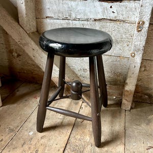 May include: A small wooden stool with a round, glossy black seat and four dark brown tapered legs. The legs are connected by a central support structure featuring a small, rounded wooden finial at the intersection of the cross-bracing.