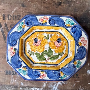 Puede incluir: Plato de cerámica octogonal con un diseño floral azul y amarillo. El plato presenta dos flores amarillas con centros rosas y hojas verdes. El borde del plato está decorado con remolinos azules y blancos.