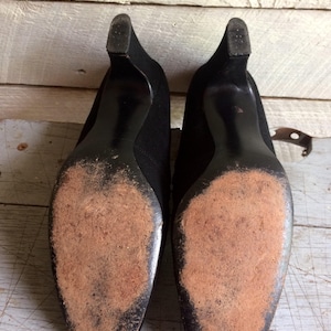 May include: A pair of black suede heeled shoes with worn leather soles. The shoes have a pointed toe and a low heel.