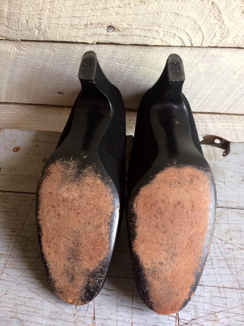 May include: A pair of black suede heeled shoes with worn leather soles. The shoes have a pointed toe and a low heel.