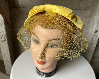 May include: A vintage yellow hat with a matching bow and a delicate net veil. The hat is displayed on a mannequin head with a black collar. The hat is a classic accessory, perfect for special occasions or vintage fashion.