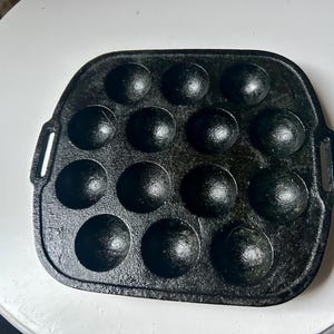 Puede incluir: Una sartén takoyaki de hierro fundido negro con 14 moldes esféricos. La sartén tiene forma rectangular con esquinas redondeadas y asas pequeñas a cada lado. La sartén está sobre una superficie blanca.